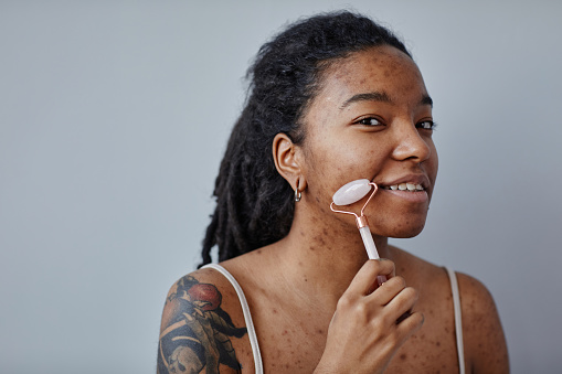 Candid portrait of young black woman with acne scars and tattoos using face massager looking at camera, copy space