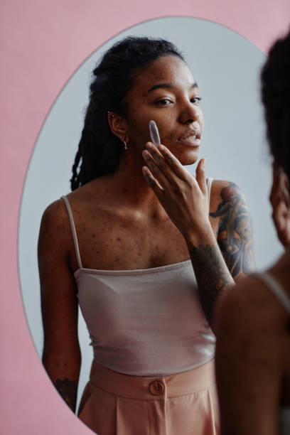 istock Young black woman with acne scars using face cream looking in mirror 1463321967