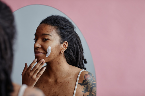 Candid portrait of young black woman with acne scars using face cream looking in mirror reflection, copy space