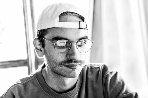 Black and white closeup portrait of 22 years old young man with baseball cap and eyeglasses using laptop, background with copy space, full frame horizontal composition