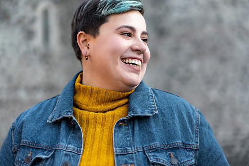 Portrait of a curvy gen z non binary woman with denim jacket and colored short hair with toothy smile looking away from camera - diversity and individuality lifestyle concept
