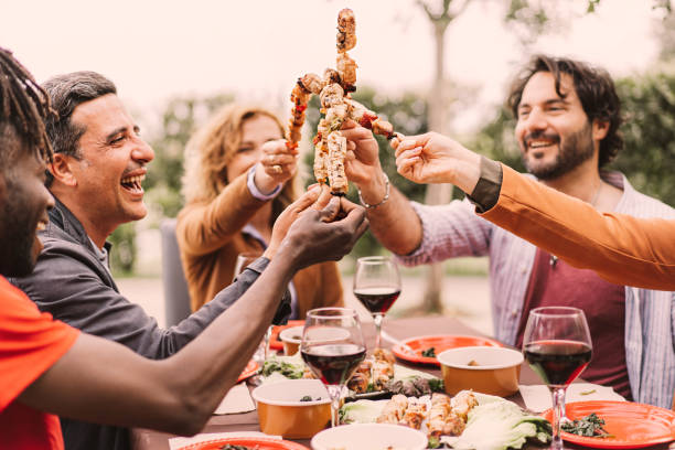 outdoor shot of multiethnic friends enjoying barbecue party in garden, sitting at the table and cheering with meat skewers. - picnic summer break relaxation imagens e fotografias de stock
