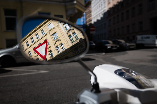 reflection of a traffic yield sign in front of a building in the side mirror of a motorcycle standing on a parking lot in a street in vienna