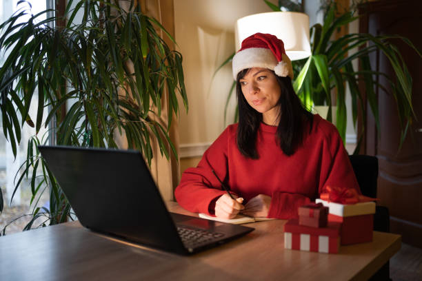 istock Cheerful joyful woman in red cap smiling toothily writing down notes at laptop desktop table on Christmas eve 1463345707