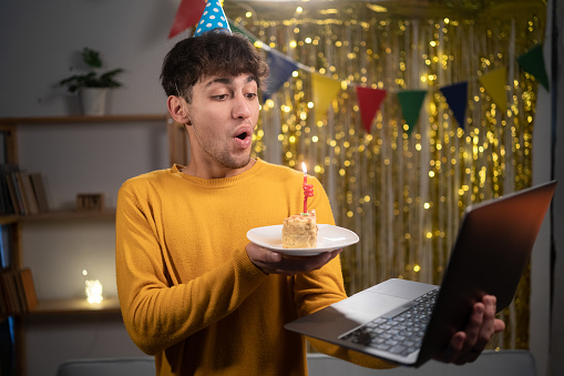Virtual celebration birthday. Happy guy in party hat celebrating birthday online, blow candle on cake, using laptop for video call with friends or family on webcam at home. Copy space