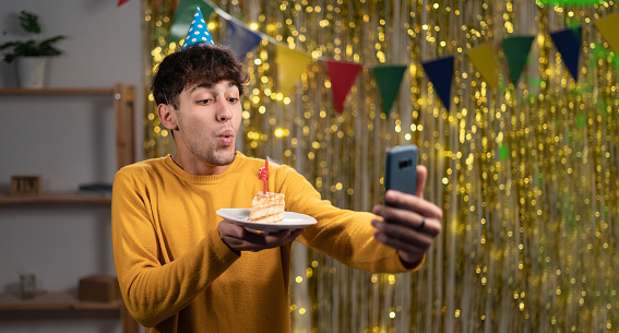 Distant congratulation. Portrait of young man holding smartphone, talking to webcam, wearing party hat, hold a piece of birthday cake and blowing a candle, celebrating virtual birthday at home. Banner