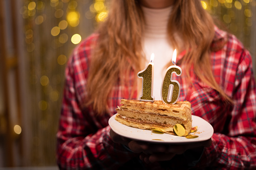 Young woman holding plate with tasty birthday cake with 16 number candle against defocused lights. Copy space