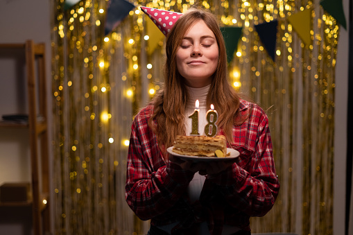 Happy 18 years old girl in party cone make a wish and blowing out candles on birthday cake, celebrate birthday at home. Copy space