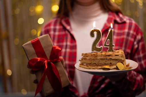 Young woman holding plate with tasty birthday cake and gift against defocused lights. 24th birthday. Copy space