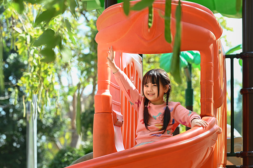 Joyful little asian girl having fun on playground in public park surrounded by green trees at sunlight morning.