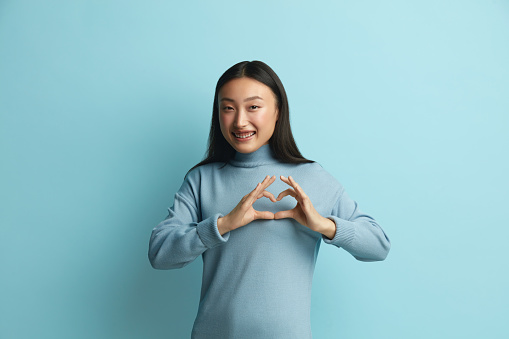 Asian Woman Showing Love Gesture. Portrait of Attractive Romantic Woman Standing Making Heart with Hands, Smiling Playfully. Indoor Studio Shot Isolated on Blue Background