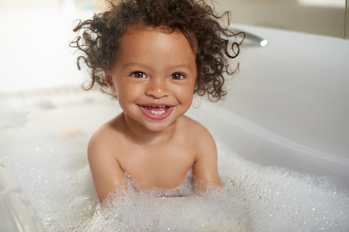 Bathtub, portrait and happy girl laughing in water, soap and foam for washing, hygiene and kids at home. Children, shower bubbles and smile in bathroom for health, wellness and fun cleaning in house