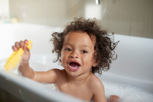 Fun, playful and portrait of a baby in the bath with toys for playing, cleaning body and routine. Happy, smile and child in bubble water for washing, bathing and clean grooming in a home bathroom