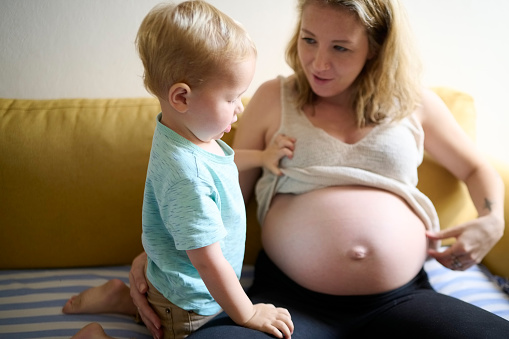 Pregnant mother with child on sofa, little boy looking at mommy's belly on sofa in living room and family at home. Pregnancy announcement with son, showing stomach and maternity growth in abdomen