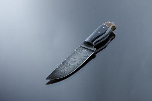 An all-metal, middle kitchen knife lies on a mirror surface, a blade made of multilayer damask steel, lining from deer antler and walnut burl