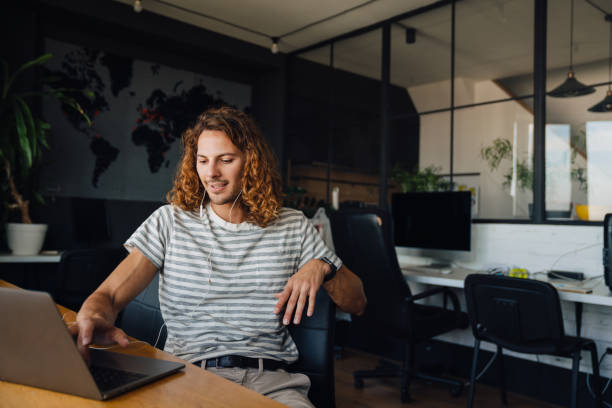 istock Young man using laptop and earphones while sitting by table in office 1463385315