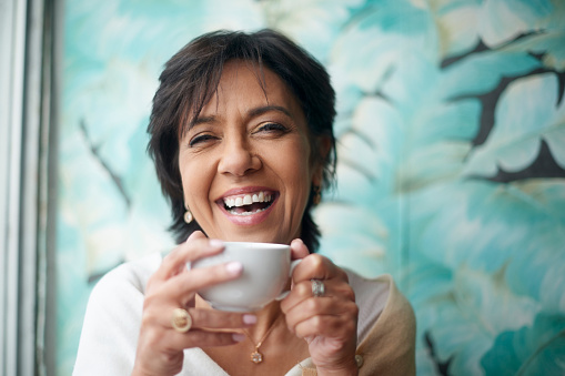Senior woman, coffee and face laughing in joy for freedom, happiness or service at an indoor cafe. Portrait of happy elderly female enjoying a warm or hot beverage drink and smiling at a restaurant