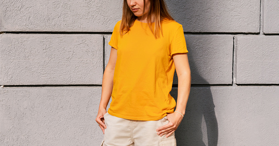 Unrecognizable young millenial blonde woman in yellow orange color T-shirt on gray wall background Mockup friendly copy space for design and text urban clothing style