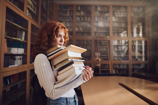 Female student holding heavy stack of books in library. Copy space.