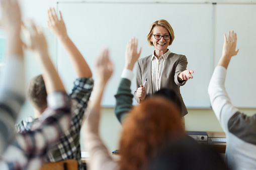 Happy mature professor aiming at her students who raised their hands to answer a question on a class at high school.