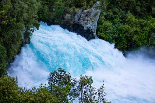 An above shot of a waterfall in Taupo in the north island of New Zealand. The water is turquoise in colour and is fast flowing. The river bank is rich in green foliage.