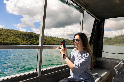 A shot of a woman taking a photo at the side of a boat in the north island of New Zealand. She is wearing sunglasses and is smiling and portraying happy emotions.