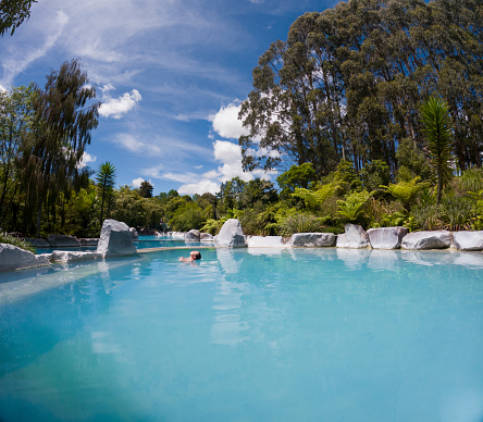 Shot of a natural geothermal pool in Taupo in the north island of New Zealand. There is a woman bathing in the pool. There is lots of green foliage and trees surrounding the pool. The water is a vibrant turquoise colour.