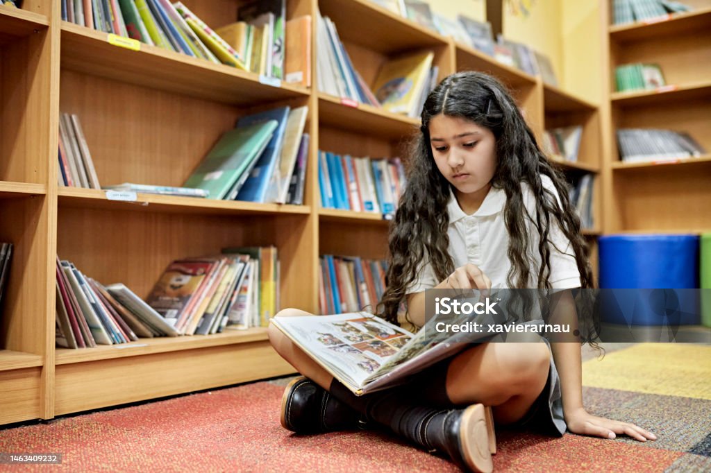 Young Hispanic student reading graphic novel Full length view of 10 year old girl in private school uniform sitting cross-legged on floor and browsing through book during visit to library. Comic Book Stock Photo