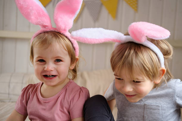 glückliche kinder, kleines mädchen und junge, tragen hasenohren stirnband spielen zu hause im schlafzimmer mit farbigen ostereiern. geschwister genießen sie die ferien. ostertag. christliches passahfest. ostern feiern. - bunny girl stock-fotos und bilder