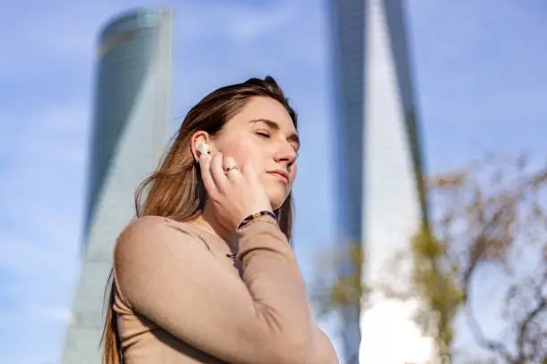 Madrid, Spain. Close-up shot of a thoughtful and concentrated beautiful young woman with her eyes closed and listening through her wireless in-ear headphones. Sunset in a financial district with skyscrapers