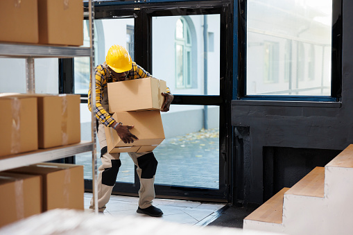 Delivery service african american loader carrying heave cardboard boxes and standing in warehouse office doors. Shipping company man employee carrying parcels in storage room