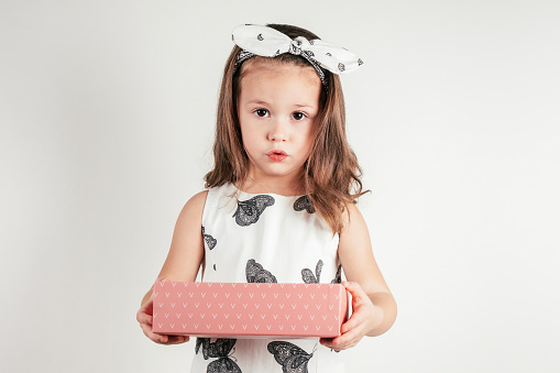 Confused little girl hold pink present box. Portrait of child on white. Pretty kid in festive dress and headband. Gift for holiday. Saint Valentine's Day concept, holiday of all lovers, celebration.