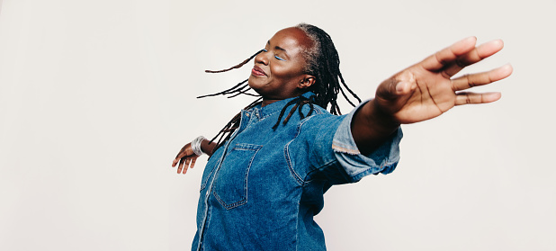 Mature happiness. Carefree woman with dreadlocks standing with her eyes closed and her arms outstretched. Cheerful middle-aged woman wearing a denim jacket and make-up in a studio.