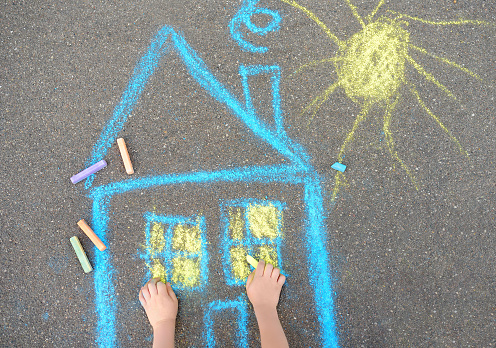 Little child boy is drawing house and sun painted with colored chalk on asphalt of sidewalk. Kids creative picture on gray background of road. Concepts of home and peaceful life all over the world.