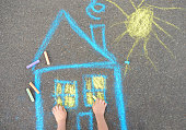 Little child boy is drawing house and sun painted with colored chalk on asphalt of sidewalk. Kids creative picture on gray background of road.