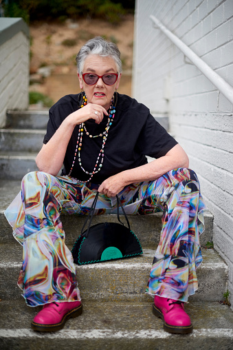 Portrait, cool and funky mature woman with stylish and trendy fashion sitting on stairs outdoors. Old, elderly and urban outfit by pensioner female with modern and colorful clothes on steps