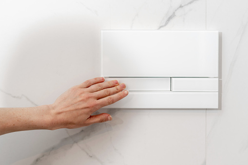 closeup of female hand pushing switch for water flush control on white marble tiled background, modern concealed cistern, detail in bathroom interior