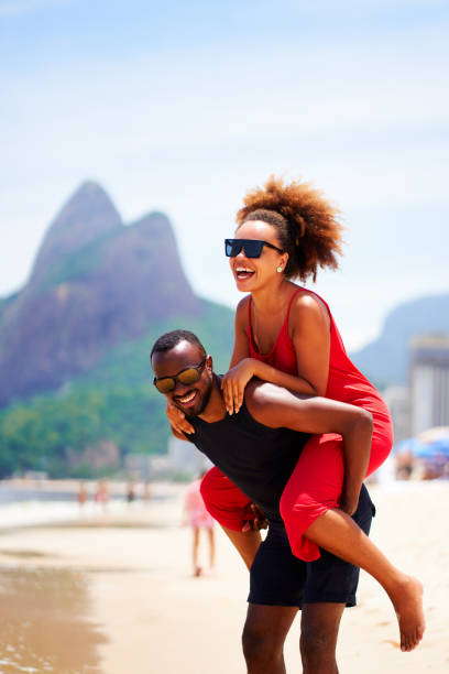junges schwarzes brasilianisches carioca-paar in huckepack-position, das lächelt und spaß am strand von ipanema hat brasilien - tourism travel travel destinations vertical stock-fotos und bilder