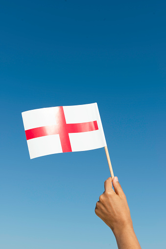 Hand is waving flag of England with clear blue sky in background.