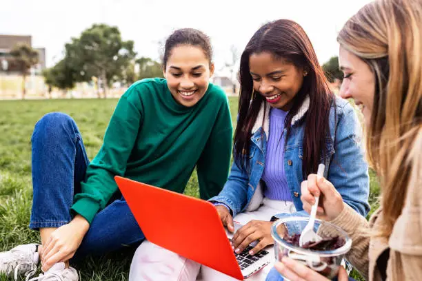 Young multiracial female students working together on laptop during lunch break sitting on green grass at college campus