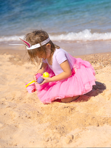 4 years old Kid on Easter egg hunt on the sandy beach. Girl with bunny ears searching for colorful eggs. Happy Easter holidays concept