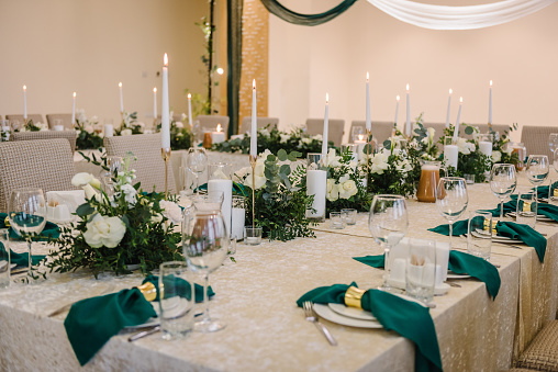 Wedding set up, dinner table reception. A plate with a green cloth towel, knives and forks next to the plate. Flower composition with eucalyptus leaves in the center of the table and burning candles.