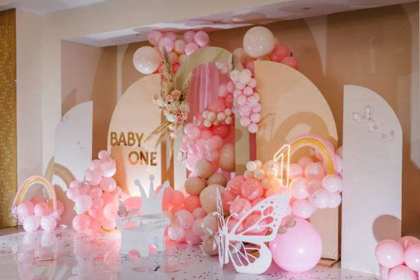 背景の写真の壁に1歳の女の子の誕生日パーティー。アーチで飾られたピンクの風船、虹、テキストの赤ちゃん、花、紙の装飾の蝶、木製の白い椅子。子供の写真ゾーン。