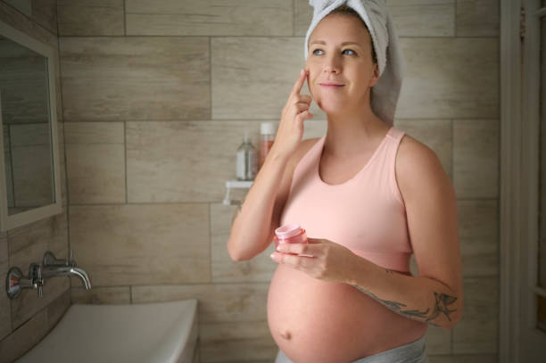 Face cream, skincare and pregnant woman in a bathroom for grooming, hygiene and facial mask. Pregnancy, beauty and girl relax with sunscreen, product and cosmetics routine, skin and luxury lotion stock photo