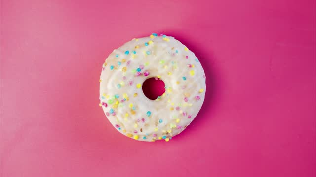 Eating delicious sweet donut with colorful sprinkles on pink background. Timelapse. Bakery and food concept. Top view of colorful frosted doughnut in 4K, UHD