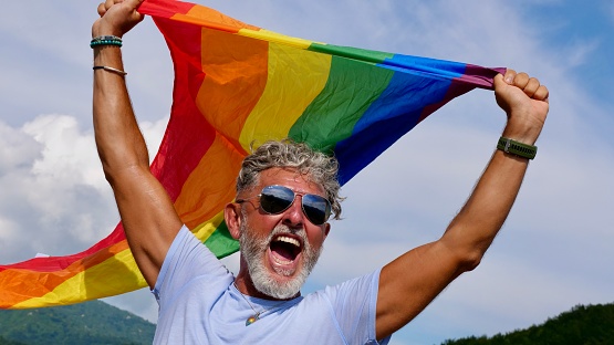 Portrait of a gray-haired elderly Caucasian man with a beard and sunglasses holding a rainbow LGBTQIA flag against a sky background, shouts in protest, Celebrates Pride Month Coming Out Day