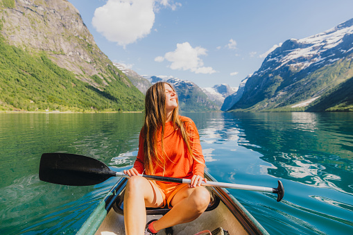 Happy female with long hair sailing in canoe on turqouise glacial lake surrounded by green mountain peaks in Scandinavia