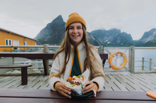 Portrait of a smiling female having outdoor dinner with seafood burger on the harbour with dramatic view of ocean and mountains in Northern Norway
