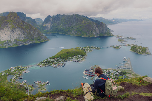 A male with backpack with a cute dog getting to the top of the mountain and text messaging with a view of the ocean and the mountain peaks on Lofoten, Northern Norway