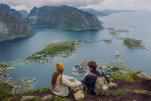 High-angle photo of happy couple with a cute dog getting to the top of the mountain and enjoying a view of the ocean and the mountain peaks on Lofoten, Northern Norway